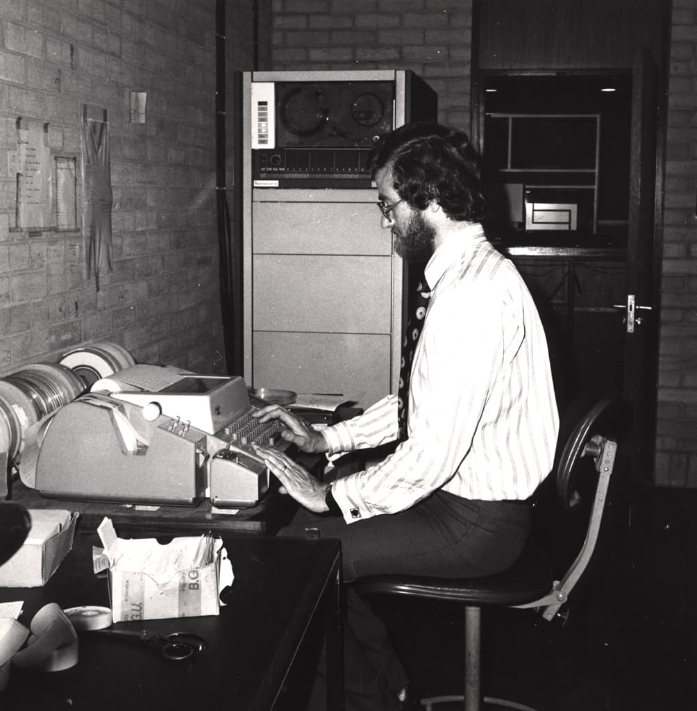 A man sitting in front of what looks like a typewriter out the top of which a sheet of paper emerges. Behind him is a big cupboard-like machine.