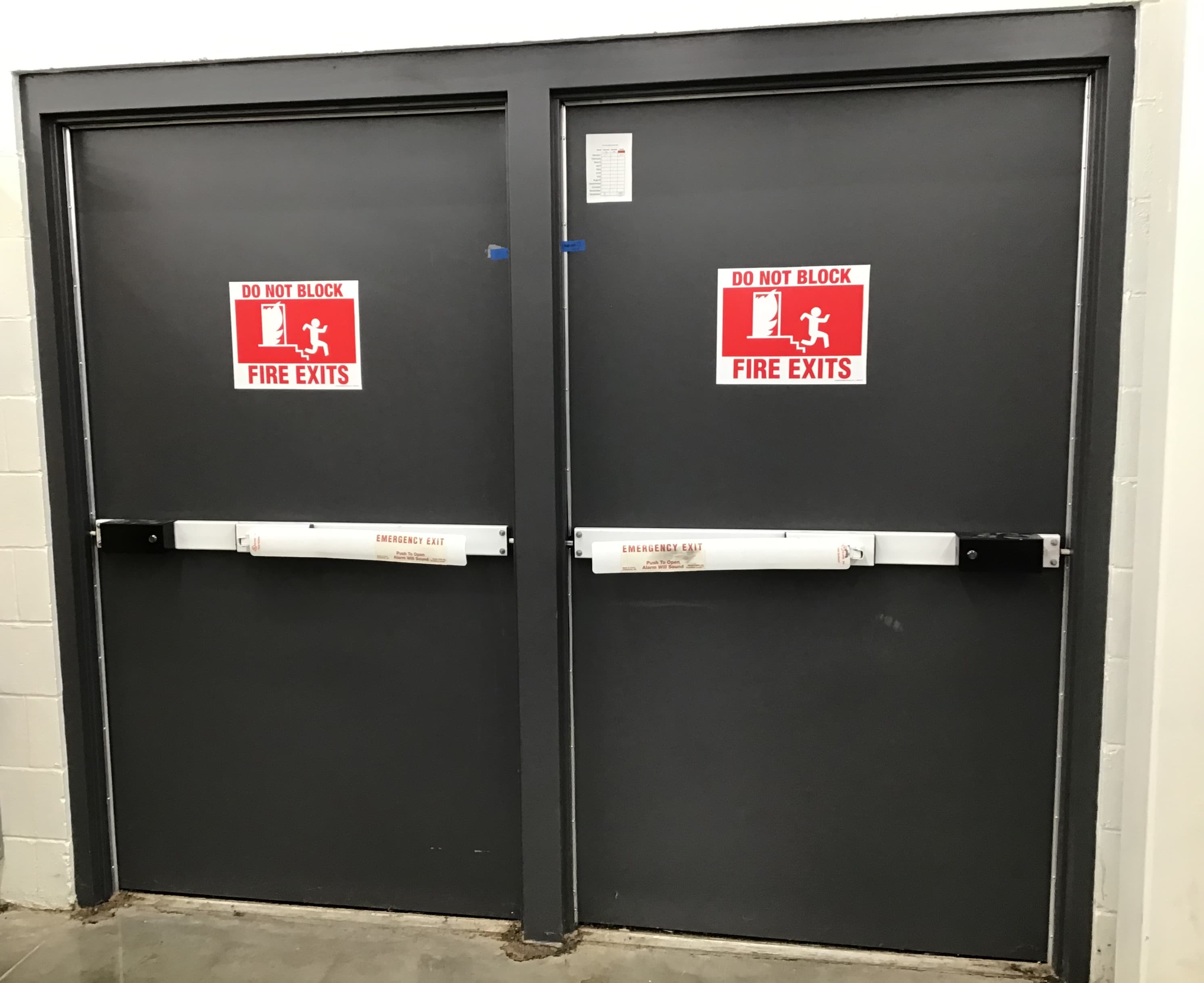 A double door with signs stating “Do not block. Fire exit.” There is a handle going across each door with a wide, pushable lever labeled "Emergency exit. Push to open. Alarm will sound."